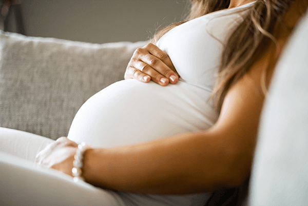 Nahaufnahme einer schwangeren Person in weißer Kleidung, die ihre Hände auf den Babybauch legt.